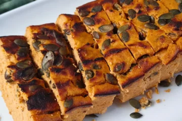 A close-up image of sliced Keto Cottage Cheese Pumpkin Bread topped with pumpkin seeds, presented on a white plate. The bread has a golden-brown crust and a soft, orange-hued interior indicative of pumpkin.