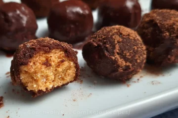 A close-up image of Keto Cottage Cheese Pumpkin Balls coated in a dark powder, displayed on a white plate with one ball cut in half to show the creamy orange filling.