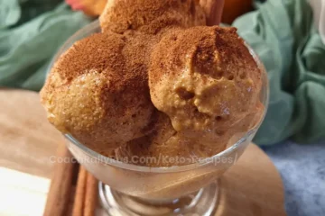 A glass bowl filled with scoops of pumpkin cottage cheese keto ice cream, sprinkled with cinnamon, presented on a wooden surface with autumn leaves and a small pumpkin in the background, illustrating a high-protein, low-carb dessert option.