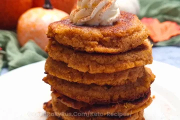A stack of six golden-brown pumpkin spice cottage cheese protein pancakes on a white plate, garnished with a dollop of cream and a sprinkle of spice on top.