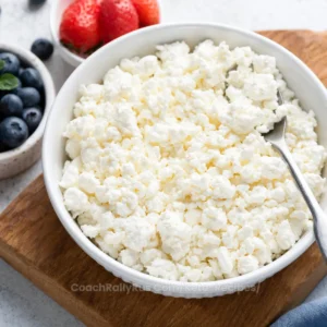A bowl of fresh cottage cheese with a spoon is placed on a wooden board. To the side, there are strawberries and blueberries, suggesting a healthy meal option. The image may be used to compare the nutritional benefits of cottage cheese versus Greek yogurt in the context of a blog post.