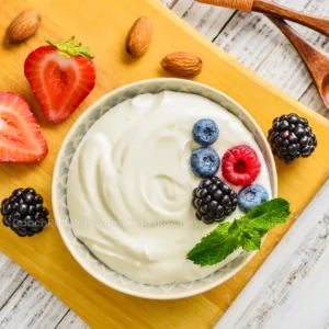 A top-down image showcasing a bowl of Greek yogurt garnished with fresh blueberries, raspberries, blackberries, a mint leaf, and accompanied by strawberries and almonds on a wooden cutting board. The creamy texture of the Greek yogurt contrasts with the vibrant colors of the berries, making it visually appealing and suggesting a healthy food choice. The image is relevant for discussions on nutritious alternatives or comparisons between cottage cheese and Greek yogurt in dietary contexts.