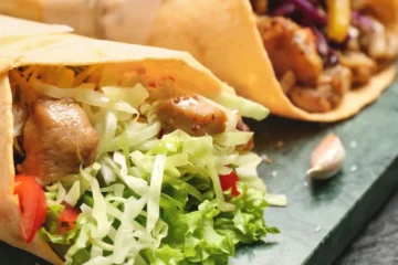 An image showcasing two types of stuffed meat wraps on a wooden surface. On the left is a Gyro with sliced meat, tomatoes, onions, and shredded lettuce wrapped in a thin flatbread. On the right is a Doner Kebab with chunks of meat, purple cabbage, onions, and greens in a thicker bread cone. Both are garnished with fresh vegetables around them.