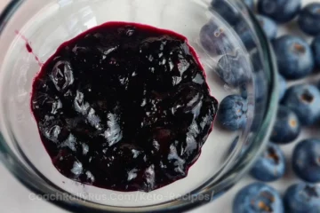 A glass jar filled with dark purple Keto blueberry jam on a white surface, with several whole blueberries scattered around it.