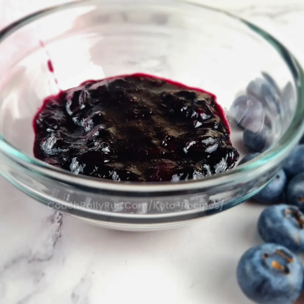 A glass jar filled with dark purple Keto blueberry jam on a white surface, with several whole blueberries scattered around it.