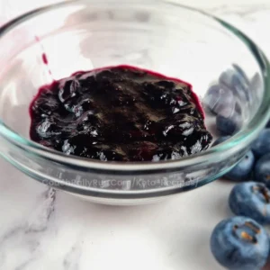 A glass jar filled with dark purple Keto blueberry jam on a white surface, with several whole blueberries scattered around it.