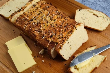 A freshly baked loaf of Keto Cottage Cheese Bread with Coconut Flour, sliced and placed on a wooden cutting board. The bread is topped with a sprinkle of seeds and herbs, giving it a rustic appearance. Beside the bread is a piece with butter on top, a block of butter, and a knife with butter residue.