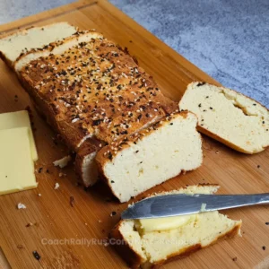 A freshly baked loaf of Keto Cottage Cheese Bread with Coconut Flour, sliced and placed on a wooden cutting board. The bread is topped with a sprinkle of seeds and herbs, giving it a rustic appearance. Beside the bread is a piece with butter on top, a block of butter, and a knife with butter residue.
