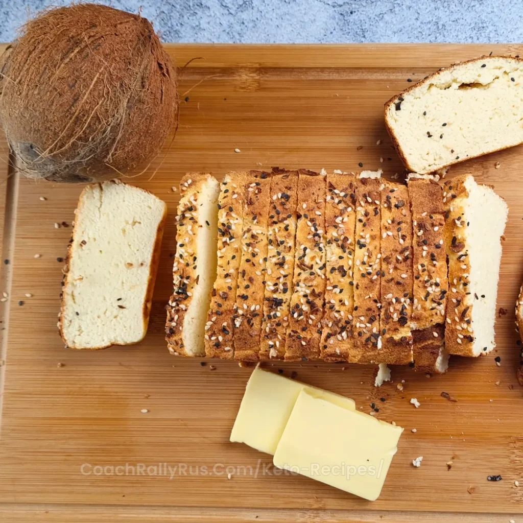 A freshly baked loaf of Keto Cottage Cheese Bread with Coconut Flour, sliced and placed on a wooden cutting board. The bread is topped with a sprinkle of seeds and herbs, giving it a rustic appearance. Beside the bread is a piece with butter on top, a block of butter, and a knife with butter residue.