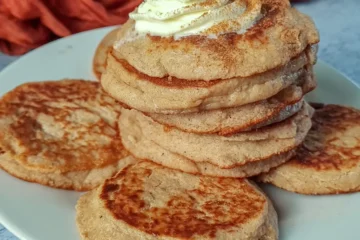 A stack of Keto Gingerbread Cottage Cheese Pancakes topped with a dollop of cream and a sprinkle of cinnamon, served on a white plate with two additional pancakes beside the stack.