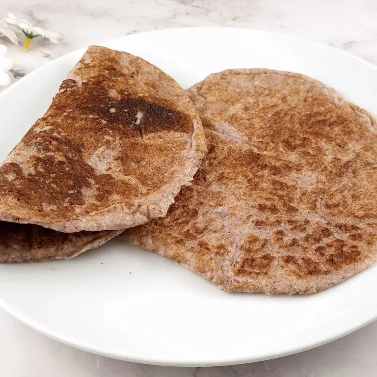 A stack of four round and thin tortillas made with almond flour and psyllium husk on a white plate. The tortillas are low-carb, gluten-free, vegan, and paleo-friendly. They have a golden brown color and a slightly cracked surface. The plate is on a wooden table with a blue napkin and a fork. The image has a text overlay that says “2 Ingredient Keto Tortillas with Almond Flour: Vegan & Paleo”