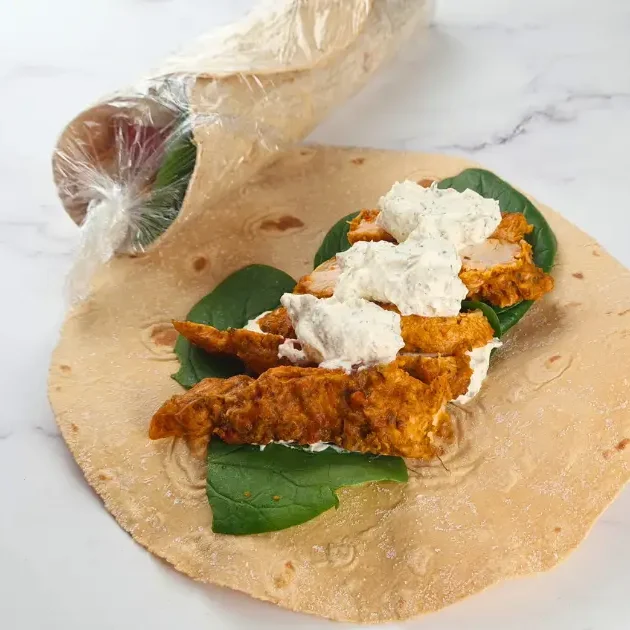 A photo of a keto chicken wrap with fried chicken and greens on a marble countertop. The wrap is open and you can see the filling, which consists of fried chicken, greens and a white sauce. The wrap is on a piece of parchment paper. The background is a white marble countertop. There is a bunch of greens and a roll of plastic wrap in the background.