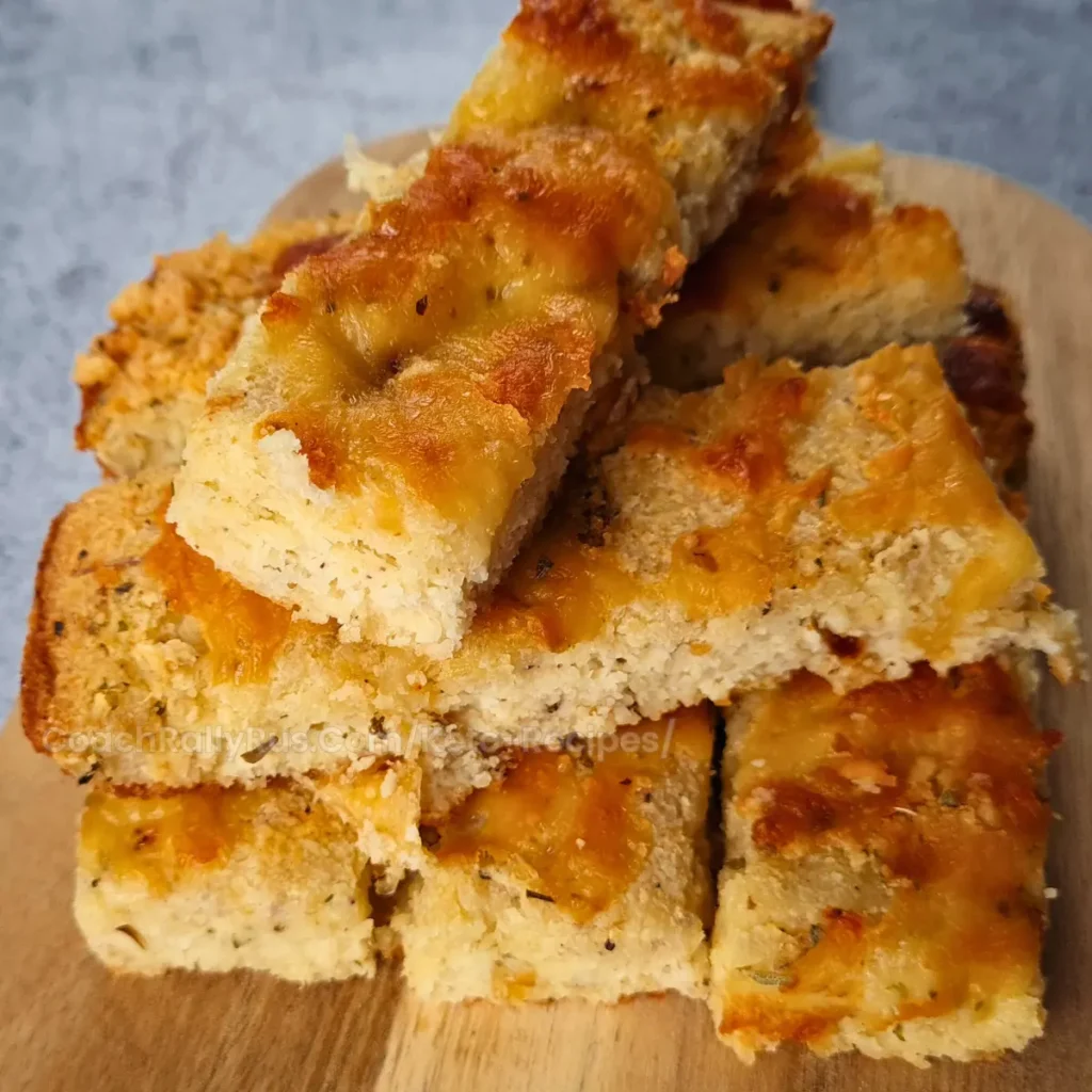 A stack of keto focaccia slices on a wooden board with melted cheese and herbs.