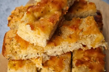 A stack of keto focaccia slices on a wooden board with melted cheese and herbs.