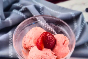 A clear glass bowl with three scoops of creamy keto strawberry crème fraîche ice cream, garnished with a fresh strawberry slice, placed on a white marble surface with a gray napkin in the background.