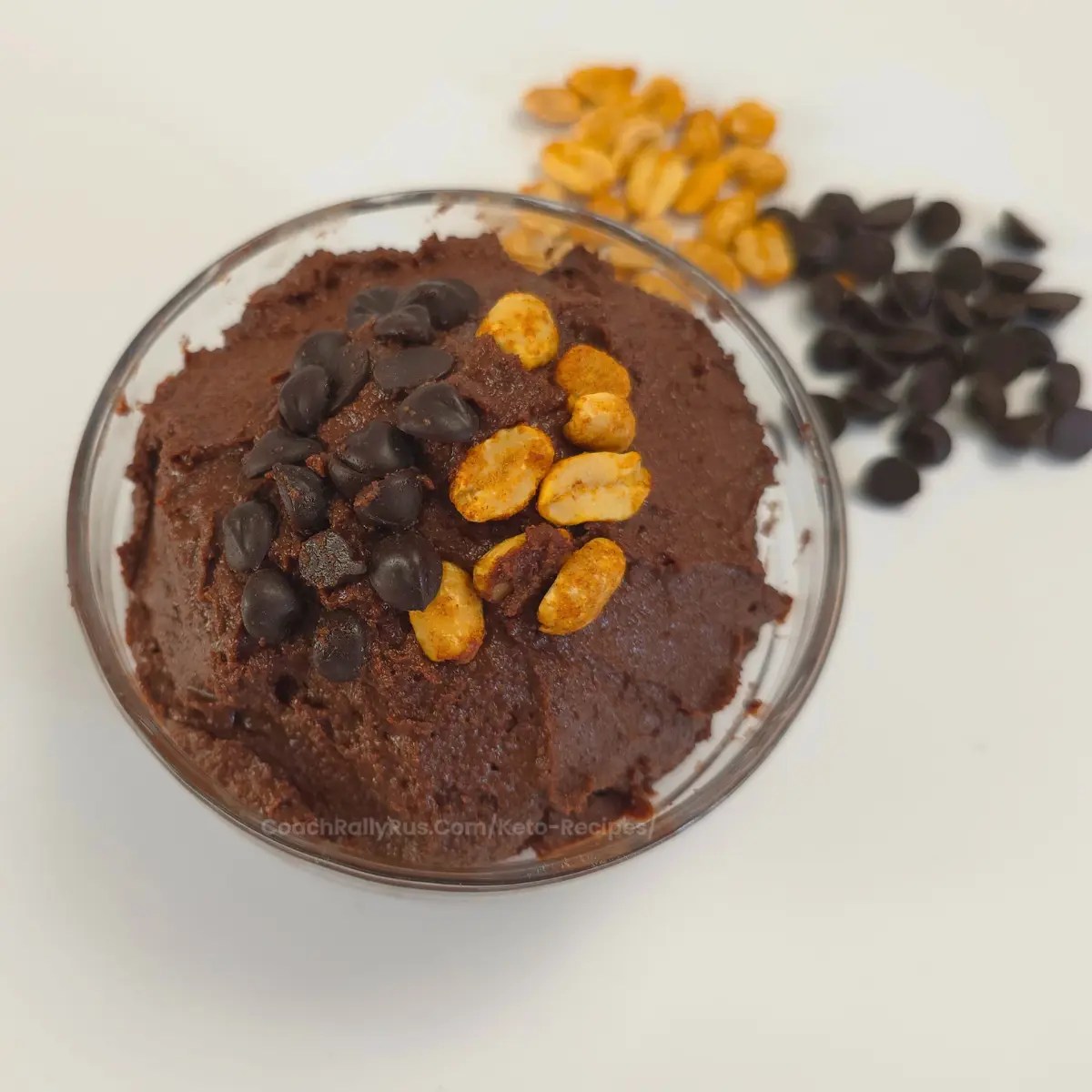 Keto-friendly chocolate peanut butter cookie dough in a glass bowl, topped with roasted peanuts and dark chocolate chips