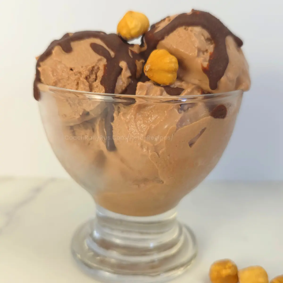 Scoops of Nutella ice cream in a glass cup topped with hazelnuts and drizzle.